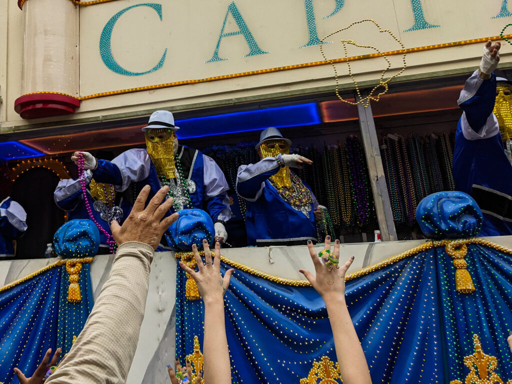 Des mains se tendent vers le flanc d'un char drapé de bleu et d'or, où des cavaliers masqués de doré, en costume bleu et chapeau blanc, lancent des colliers de perles ; des centaines de colliers pendent derrière eux