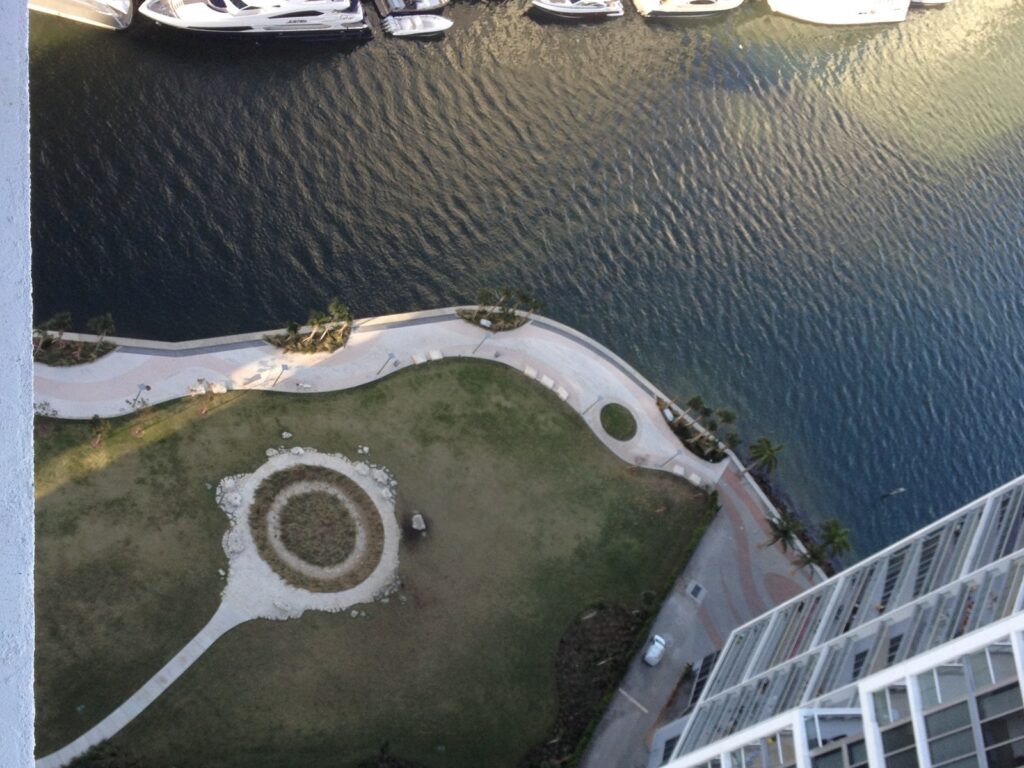 Vue plongeante depuis une tour de Brickell : sur une pelouse au bord de la rivière Miami, un cercle de pierres et de végétation marque l'emplacement du site tequesta du Miami Circle, avec des yachts amarrés sur l'autre rive