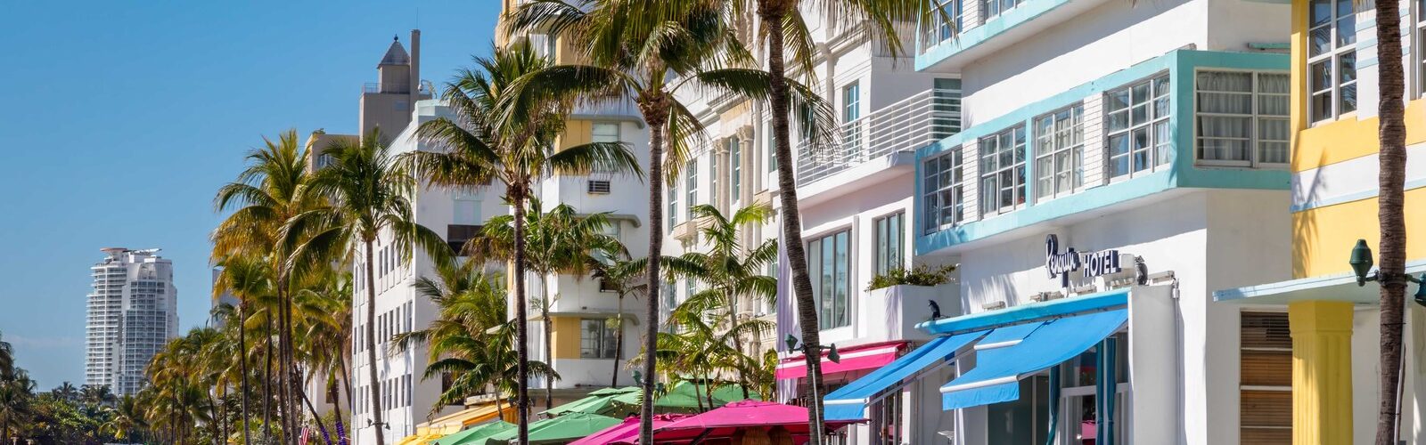 Ocean Drive à South Beach sous un ciel bleu : façades Art déco blanches, jaunes et bleu pâle, palmiers, terrasses aux parasols colorés et piste cyclable verte le long de l'avenue