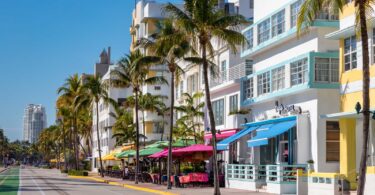 Ocean Drive à South Beach sous un ciel bleu : façades Art déco blanches, jaunes et bleu pâle, palmiers, terrasses aux parasols colorés et piste cyclable verte le long de l'avenue