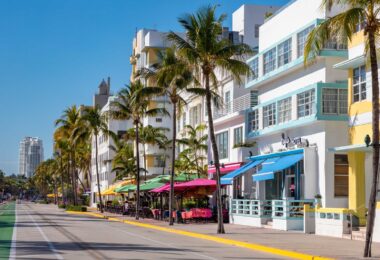 Ocean Drive à South Beach sous un ciel bleu : façades Art déco blanches, jaunes et bleu pâle, palmiers, terrasses aux parasols colorés et piste cyclable verte le long de l'avenue