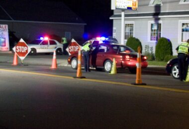 De nuit, des policiers en gilet jaune contrôlent des voitures entre des cônes orange et des panneaux Stop, devant des voitures de police aux gyrophares allumés