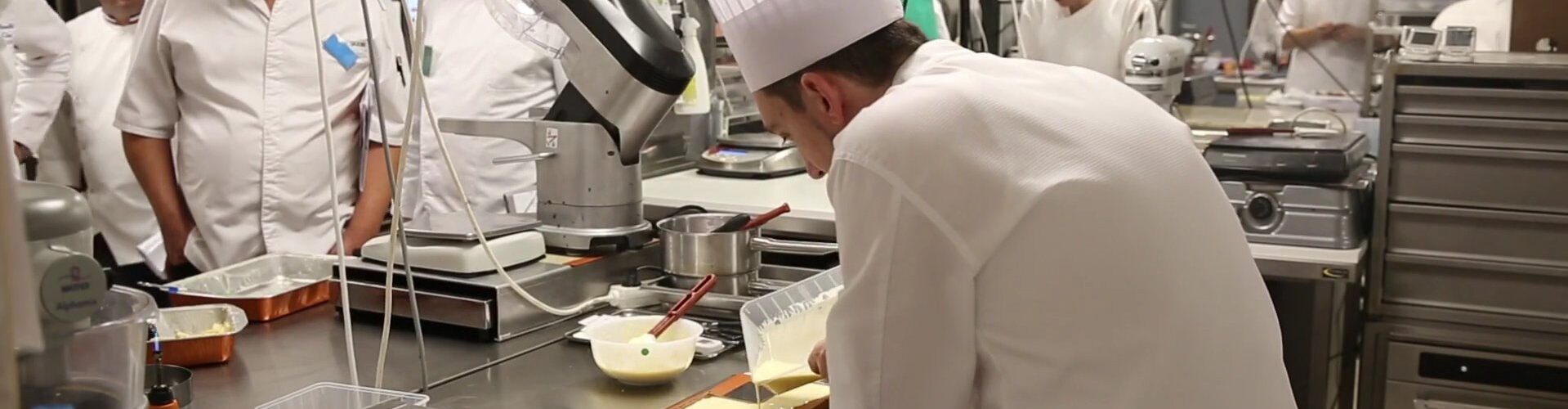 Un candidat en toque travaille une préparation pendant l’épreuve de pâtisserie du concours Meilleur Ouvrier de France, observé par des membres du jury