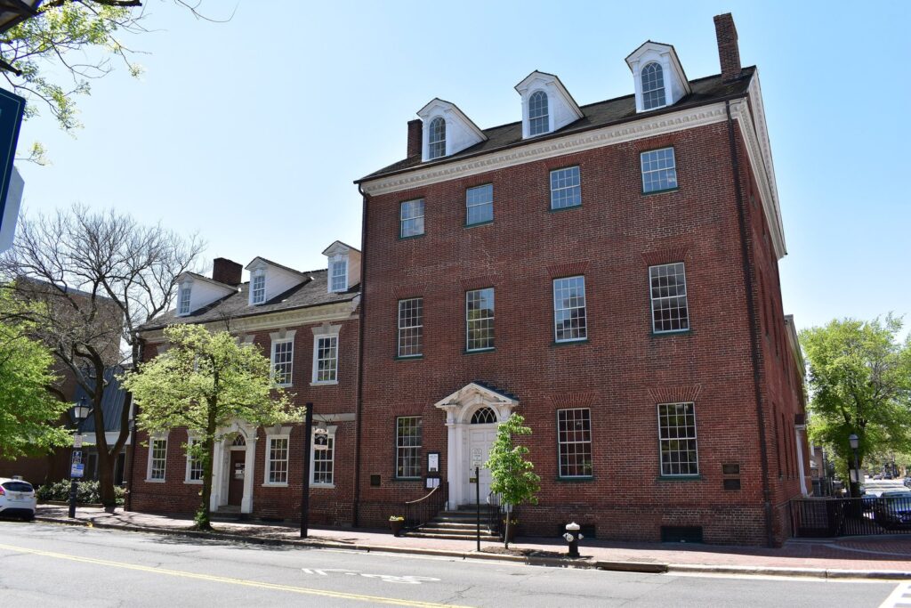 Façade en brique rouge de Gadsby's Tavern, bâtiment du XVIIIe siècle à lucarnes et porte à fronton blanc, au coin d'une rue d'Old Town Alexandria sous un ciel bleu