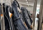Gros plan sur une longue rangée de jeans d'occasion suspendus à des cintres transparents, avec leurs étiquettes de prix, dans l'allée d'une grande friperie aux néons