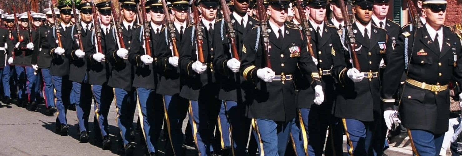 Des soldats de l'armée américaine en grand uniforme bleu, gants blancs et fusils à l'épaule, défilent en rangs serrés dans une rue d'Old Town Alexandria bordée de maisons de brique et de bois, pendant la parade de l'anniversaire de George Washington