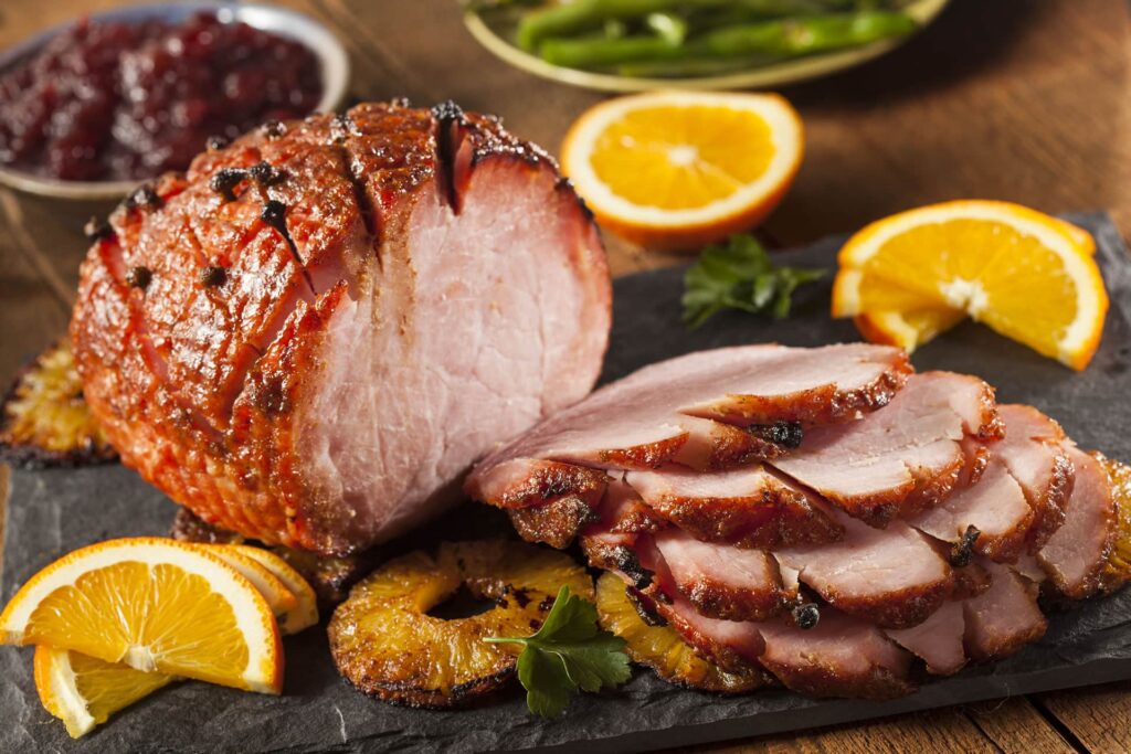 Jambon glacé au miel, piqué de clous de girofle, en partie tranché sur une ardoise, avec des rondelles d'orange et d'ananas, de la sauce aux canneberges et des haricots verts à l'arrière-plan