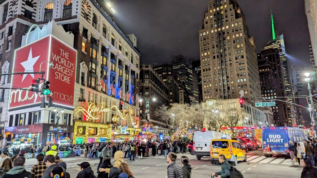Façade du grand magasin Macy's la nuit, enseigne rouge « The World's Largest Store », lettres lumineuses « Give Love » au-dessus des vitrines, foule sur le trottoir et taxi jaune sur la 34th Street