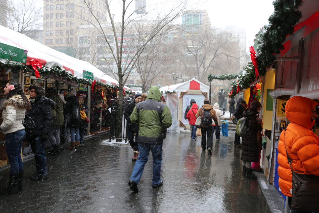 Allée d'un marché de Noël sous la neige, bordée de chalets à toit rayé rouge et blanc décorés de guirlandes, avec des visiteurs en manteau