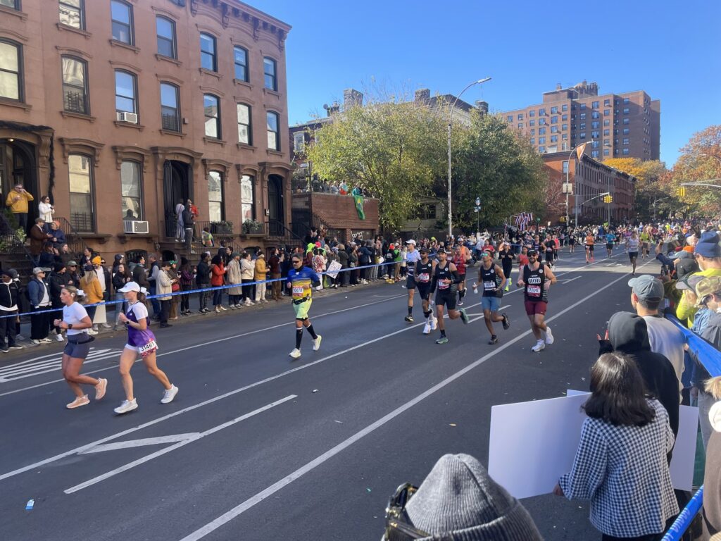 Des coureurs du marathon de New York sur une large avenue de Brooklyn bordée de maisons de brique brune, devant une foule de spectateurs massés derrière les barrières, sous un ciel bleu d'automne