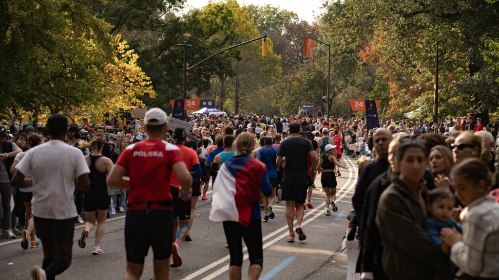 Des coureurs de dos dans une allée de Central Park aux arbres d'automne, entre deux haies de spectateurs, près du panneau orange du 24e mile