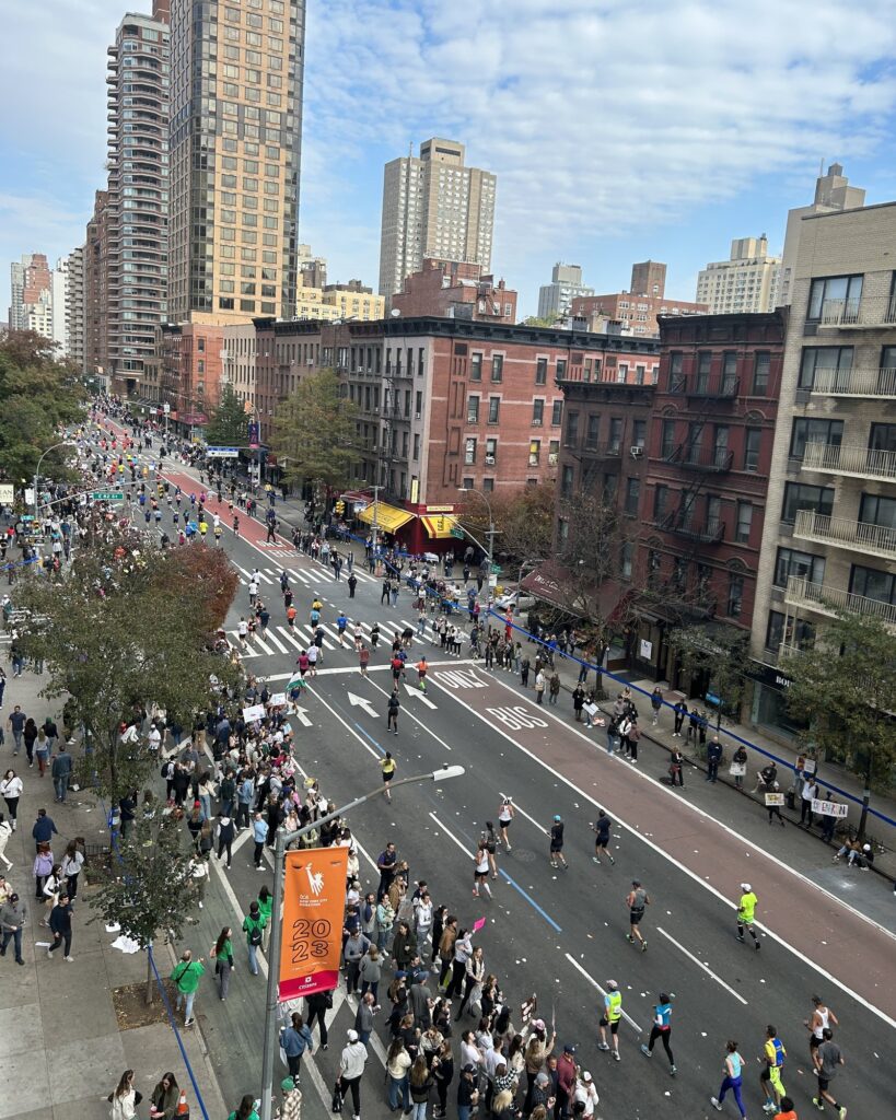 Vue plongeante depuis un balcon sur la First Avenue, dans l'Upper East Side : les coureurs remontent l'avenue entre deux foules de spectateurs, sous une bannière orange TCS New York City Marathon 2023