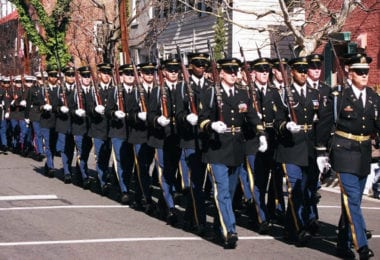 Des soldats de l'armée américaine en grand uniforme bleu, gants blancs et fusils à l'épaule, défilent en rangs serrés dans une rue d'Old Town Alexandria bordée de maisons de brique et de bois, pendant la parade de l'anniversaire de George Washington
