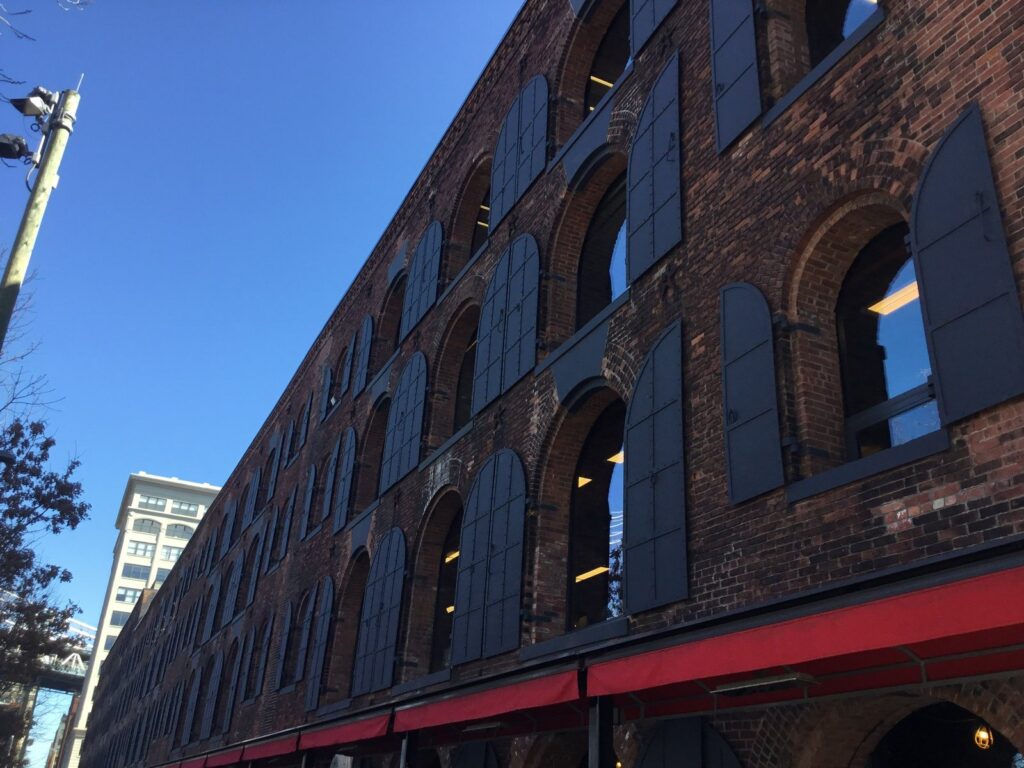 Façade d'un ancien entrepôt en brique à arcades et volets de métal, auvents rouges au rez-de-chaussée, pont suspendu visible à gauche