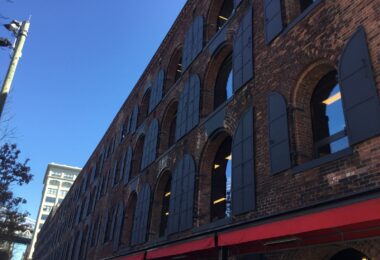 Façade d'un ancien entrepôt en brique à arcades et volets de métal, auvents rouges au rez-de-chaussée, pont suspendu visible à gauche