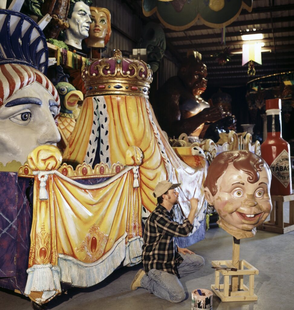 Dans un grand entrepôt, un artisan à genoux peint un char orné d'une couronne et de drapés dorés, entouré de têtes géantes, d'un visage d'enfant souriant sur un socle et d'une bouteille géante de sauce piquante