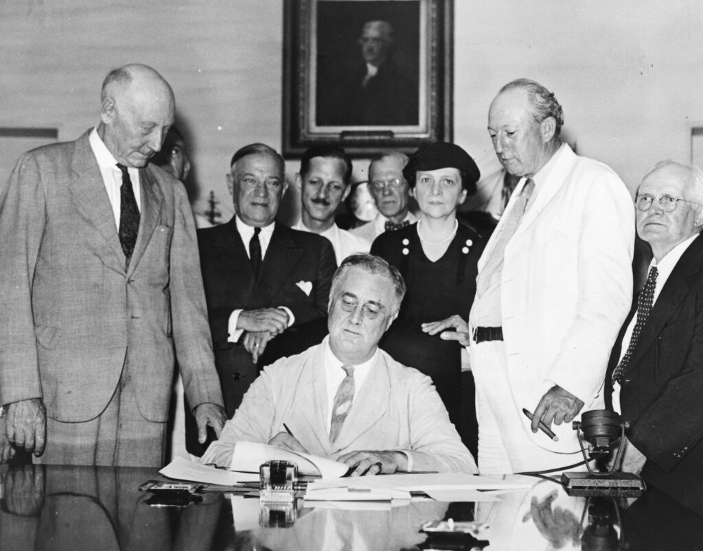Photo en noir et blanc : Franklin Roosevelt, assis à une table, signe le Social Security Act entouré de parlementaires et de la ministre du Travail Frances Perkins
