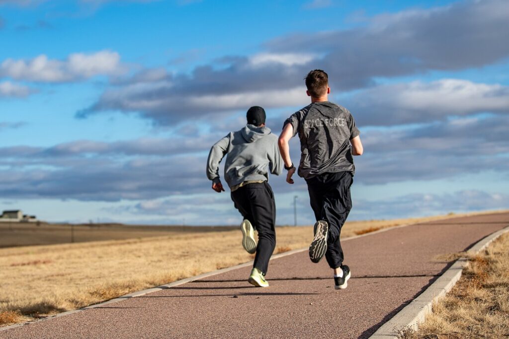 Deux coureurs sur un chemin goudronné bordé d'herbes sèches, sous un ciel bleu nuageux