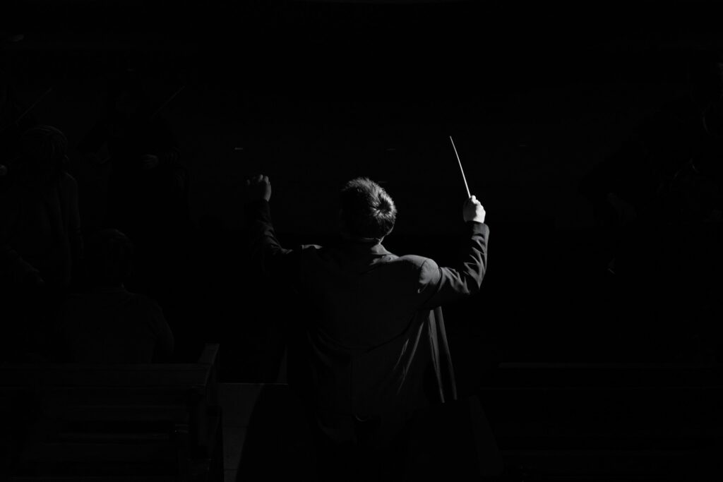Silhouette en noir et blanc d'un chef d'orchestre vu de dos, bras levés et baguette à la main, devant des musiciens dans la pénombre