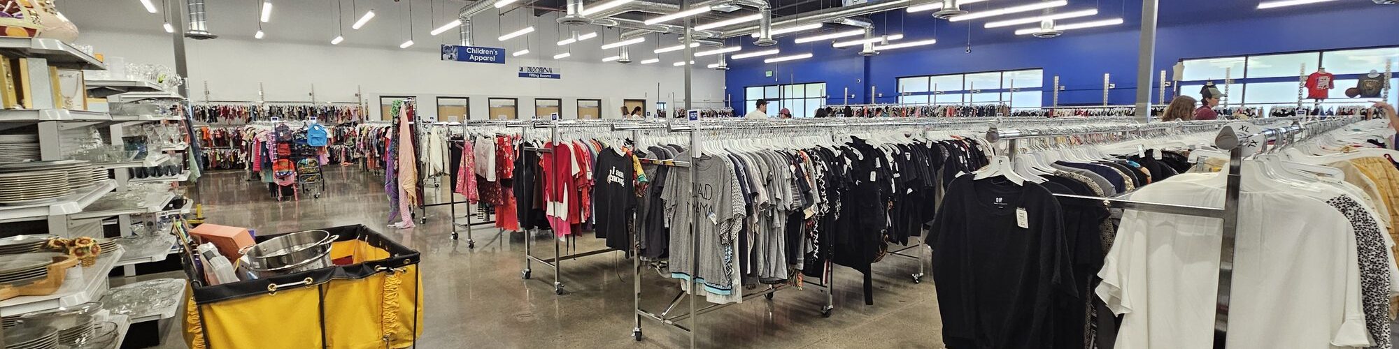 Grand magasin Goodwill aux plafonds noirs et néons : longues rangées de portants de vêtements, panneau bleu « Ladies’ Apparel », étagères de vaisselle et de verres à gauche, un chariot de tri jaune au premier plan