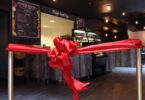 Ruban de satin rouge noué entre deux poteaux dorés devant le comptoir et la vitrine d’un café, juste avant son inauguration