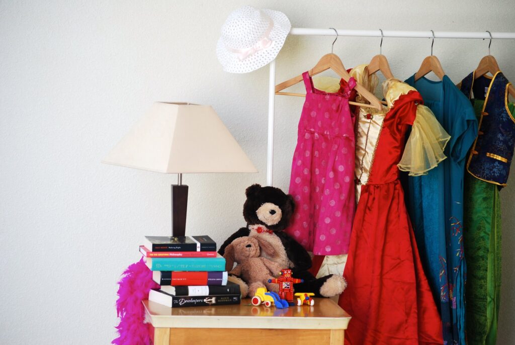 Lampe à abat-jour blanc, pile de livres, ours en peluche et petits jouets sur une commode, à côté d'un portant de robes d'enfant roses, rouges, bleues et vertes, sur fond de mur blanc