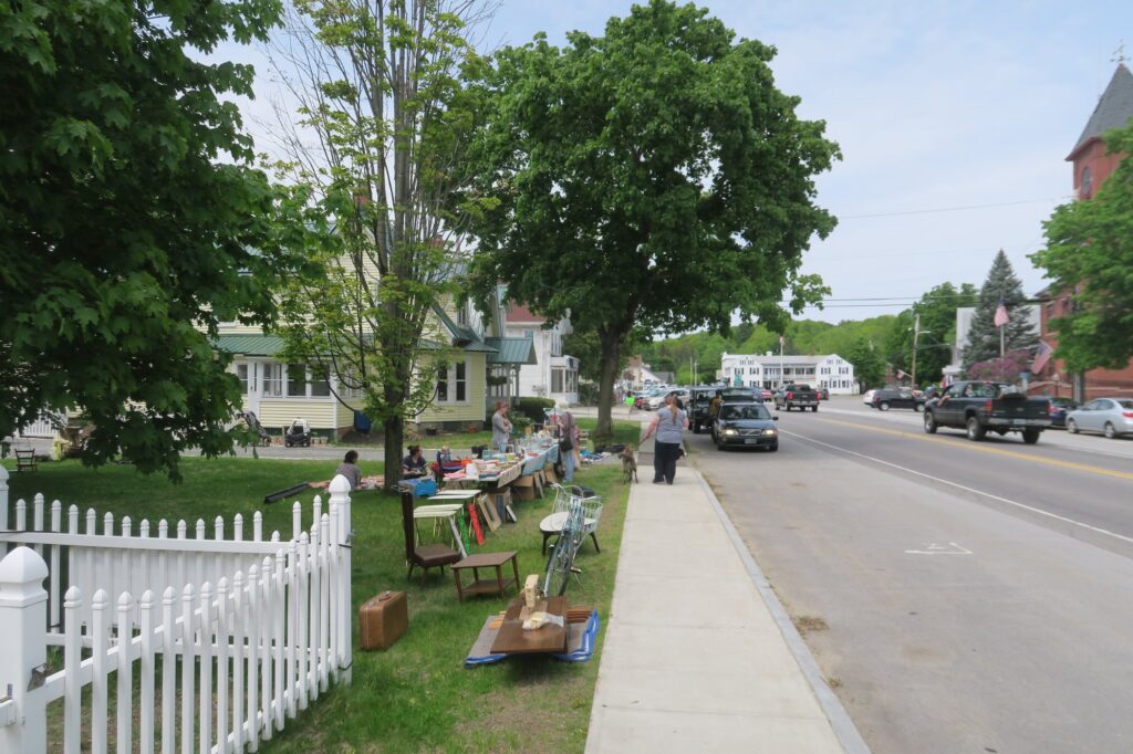 Chaise, petite table, valise, vélo, cadres et une longue table de vaisselle posés sur la pelouse derrière une barrière blanche, le long d'une rue de petite ville ; des vendeurs assis dans l'herbe et une passante avec son chien sur le trottoir