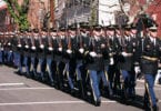 Des soldats de l'armée américaine en grand uniforme bleu, gants blancs et fusils à l'épaule, défilent en rangs serrés dans une rue d'Old Town Alexandria bordée de maisons de brique et de bois, pendant la parade de l'anniversaire de George Washington