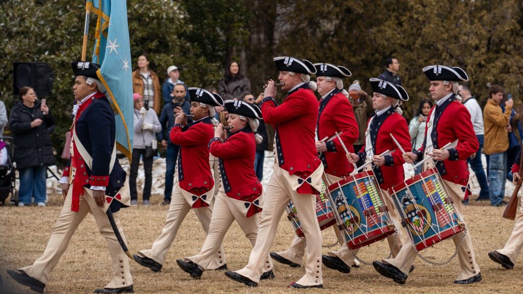 Des musiciens en habit rouge et tricorne jouent du fifre et du tambour en défilant sur une pelouse devant des spectateurs