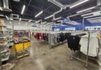 Grand magasin Goodwill aux plafonds noirs et néons : longues rangées de portants de vêtements, panneau bleu « Ladies’ Apparel », étagères de vaisselle et de verres à gauche, un chariot de tri jaune au premier plan