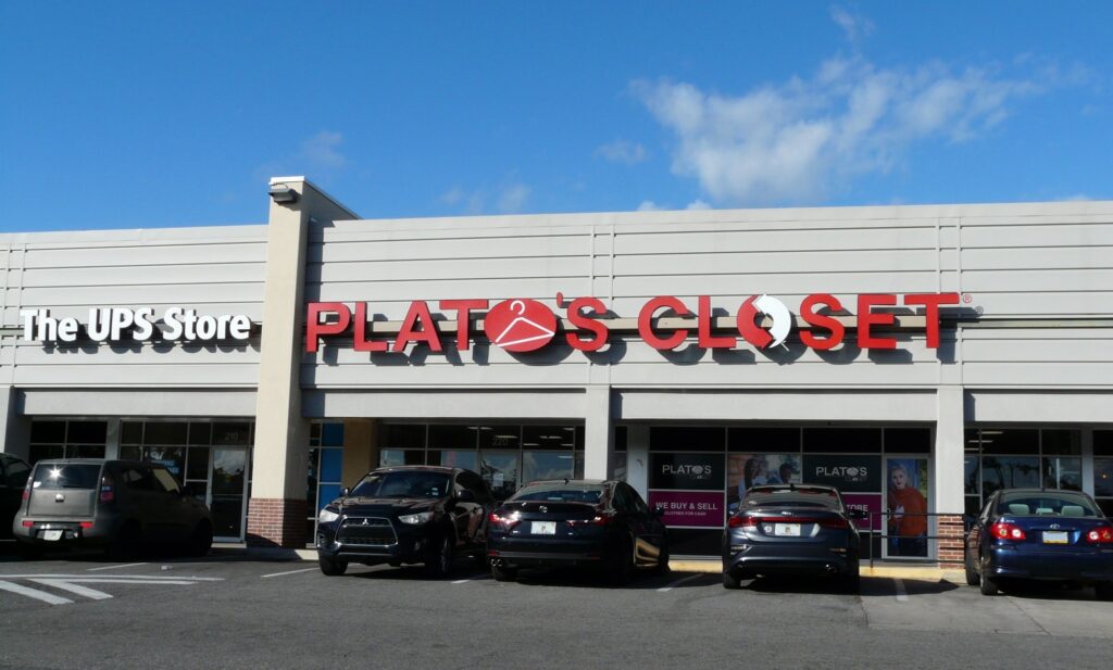 Enseigne rouge Plato's Closet sur un centre commercial de plain-pied, vitrines marquées We buy and sell, voitures garées devant, ciel bleu