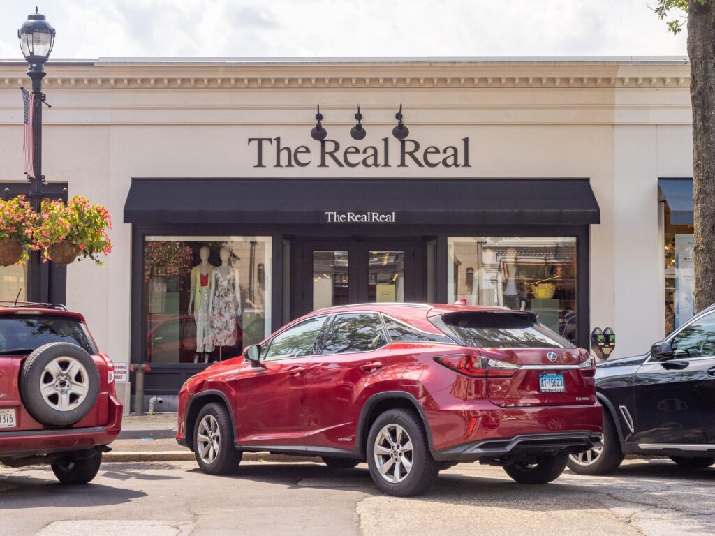 Façade crème d'une boutique au nom de The RealReal, auvent noir, vitrine avec deux mannequins en robe d'été, une voiture rouge garée devant, sur une rue commerçante