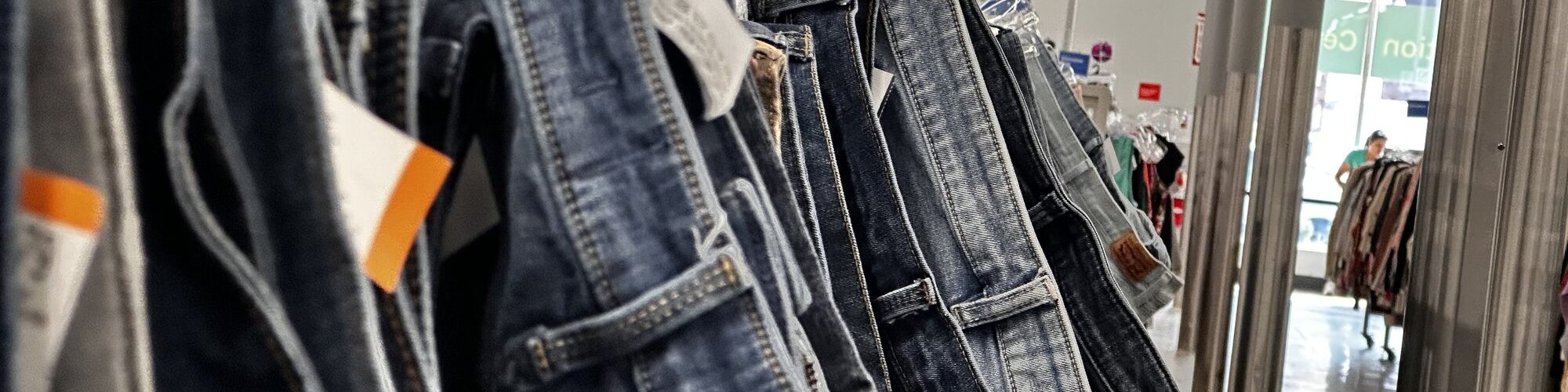 Gros plan sur une longue rangée de jeans d'occasion suspendus à des cintres transparents, avec leurs étiquettes de prix, dans l'allée d'une grande friperie aux néons