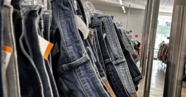 Gros plan sur une longue rangée de jeans d'occasion suspendus à des cintres transparents, avec leurs étiquettes de prix, dans l'allée d'une grande friperie aux néons