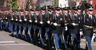 Des soldats de l'armée américaine en grand uniforme bleu, gants blancs et fusils à l'épaule, défilent en rangs serrés dans une rue d'Old Town Alexandria bordée de maisons de brique et de bois, pendant la parade de l'anniversaire de George Washington