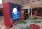 Décor The Elf on the Shelf dans une galerie marchande : grande fenêtre de nuit avec la lune, lutins en costume rouge et sapin décoré
