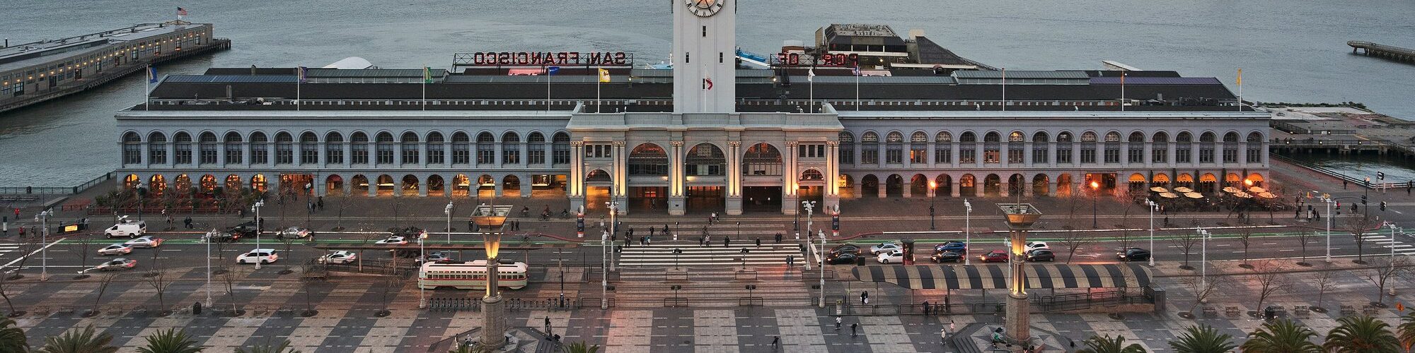 Vue en hauteur du Ferry Building de San Francisco au crépuscule, tour de l'horloge illuminée, Embarcadero bordé de palmiers, baie et Bay Bridge en arrière-plan