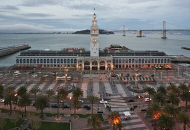 Vue en hauteur du Ferry Building de San Francisco au crépuscule, tour de l'horloge illuminée, Embarcadero bordé de palmiers, baie et Bay Bridge en arrière-plan