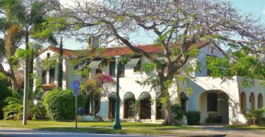 Maison blanche de style méditerranéen à toit de tuiles rouges, stores rayés verts et arcades, sous un grand arbre et des palmiers, dans une rue de Miami Shores