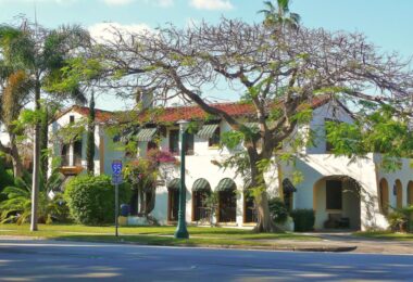 Maison blanche de style méditerranéen à toit de tuiles rouges, stores rayés verts et arcades, sous un grand arbre et des palmiers, dans une rue de Miami Shores