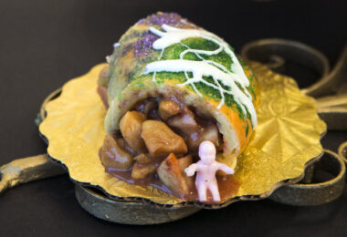 Part de king cake roulée, glacée de blanc et saupoudrée de sucre violet, vert et or, à la garniture en gros morceaux, avec une petite figurine de bébé en plastique rose posée devant, sur un carton doré