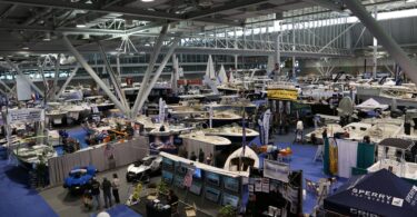 Vue en hauteur sur un hall d'exposition du New England Boat Show, avec des dizaines de bateaux à moteur et de voiliers sous une charpente métallique blanche et des stands sur une moquette bleue