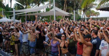 Une foule en maillot de bain et en tenue d'été lève les bras sous des voiles d'ombrage blanches et des palmiers, au bord d'une piscine, pendant une pool party de la Winter Music Conference à Miami